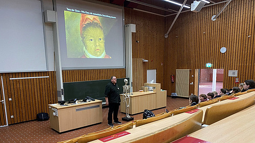Ein Mann steht in einem Hörsaal, neben ihm steht ein menschliches Skelett.