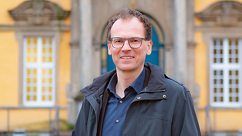 Prof. Woppowa steht im Innenhof des Osnabr&uuml;cker Schlosses. Er l&auml;chelt in die Kamera. In der Hand h&auml;lt er eine rote Mappe mit dem Logo der Universit&auml;t Osnabr&uuml;ck