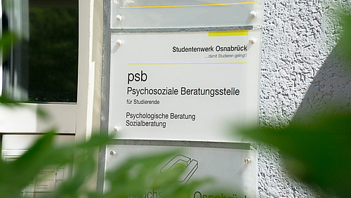 Das Bild zeigt an der Fassade eines Gebäudes ein Schild mit der Aufschrift "psb Psychosoziale Beratungsstelle für Studierende" sowie "Psychologische Beratung Sozialberatung". Im Vordergrund sind Äste mit grünen Blättern zu erkennen.