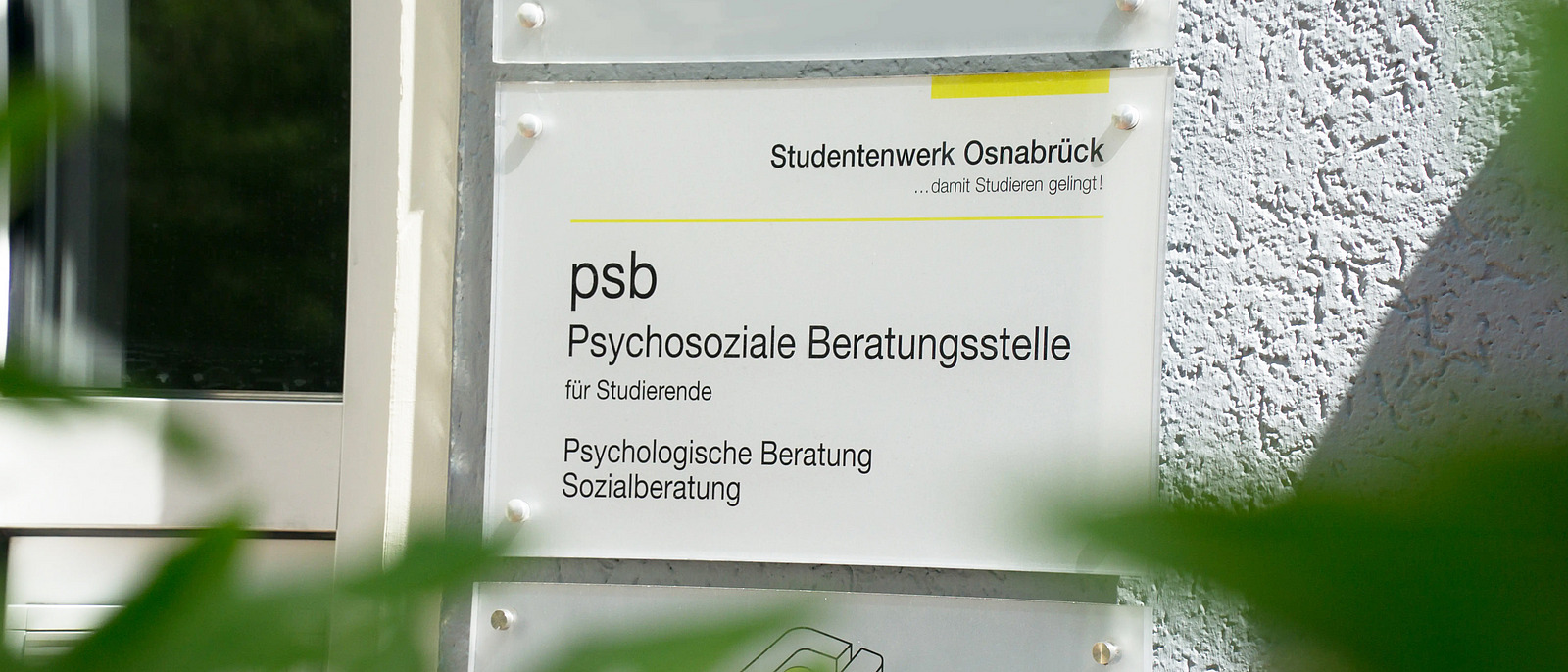 The picture shows a sign on the facade of a building with the words “psb Psychosocial Counseling Center for Students” and “Psychological Counseling Social Counseling.” Branches with green leaves can be seen in the foreground.