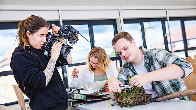 A young woman dressed in black uses a large camera to photograph a piece of excavated meadow, which is being positioned on a table in front of her by a man. In the background, another woman is reading a file folder.