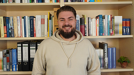 Ein  Mann mit Bart und kurzen dunklen Haaren steht lächelnd vor einem Bücherregal. Er trägt einen beigen Hoodie.