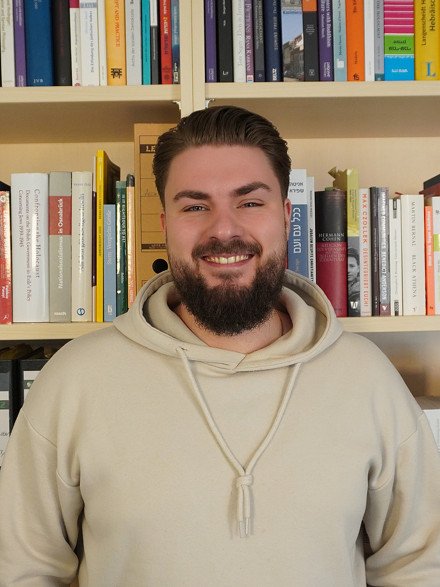 Ein  Mann mit Bart und kurzen dunklen Haaren steht lächelnd vor einem Bücherregal. Er trägt einen beigen Hoodie.
