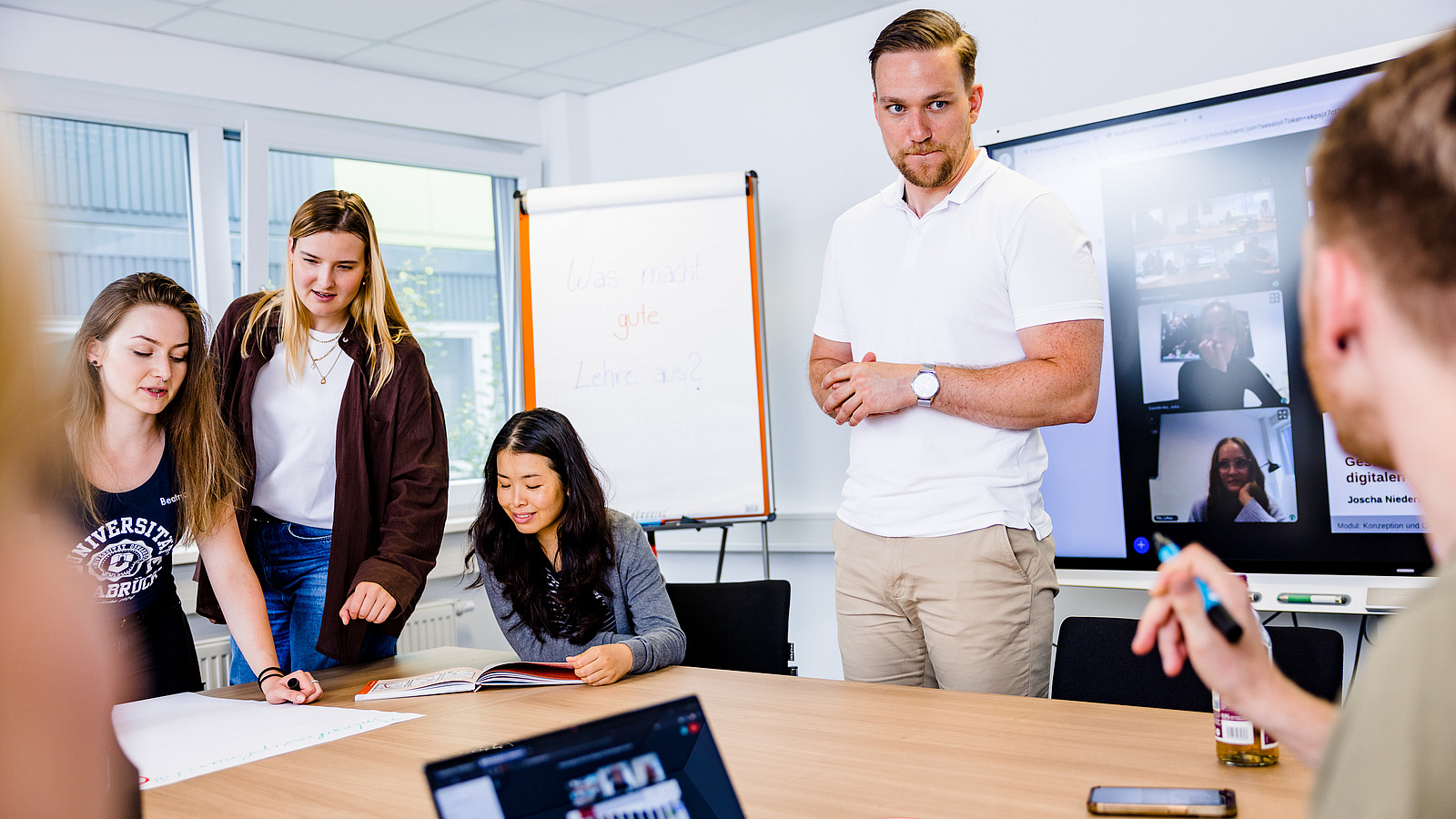 Mehrere Studierende arbeiten in einem Seminarraum. Im Hintergrund ist ein Bildschirm zu sehen, auf dem weitere Studierende in einer Videokonferenz zugeschaltet sind.
