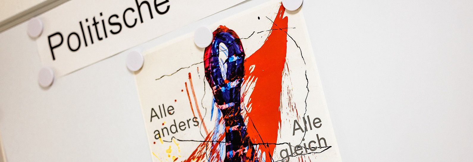 Whiteboard mit Magneten, auf einem angehefteten Zettel steht "Politische Theorie", darunter ist ein Plakat mit bunten Farbspritzern befestigt, auf dem "alle anders - alle gleich" steht.