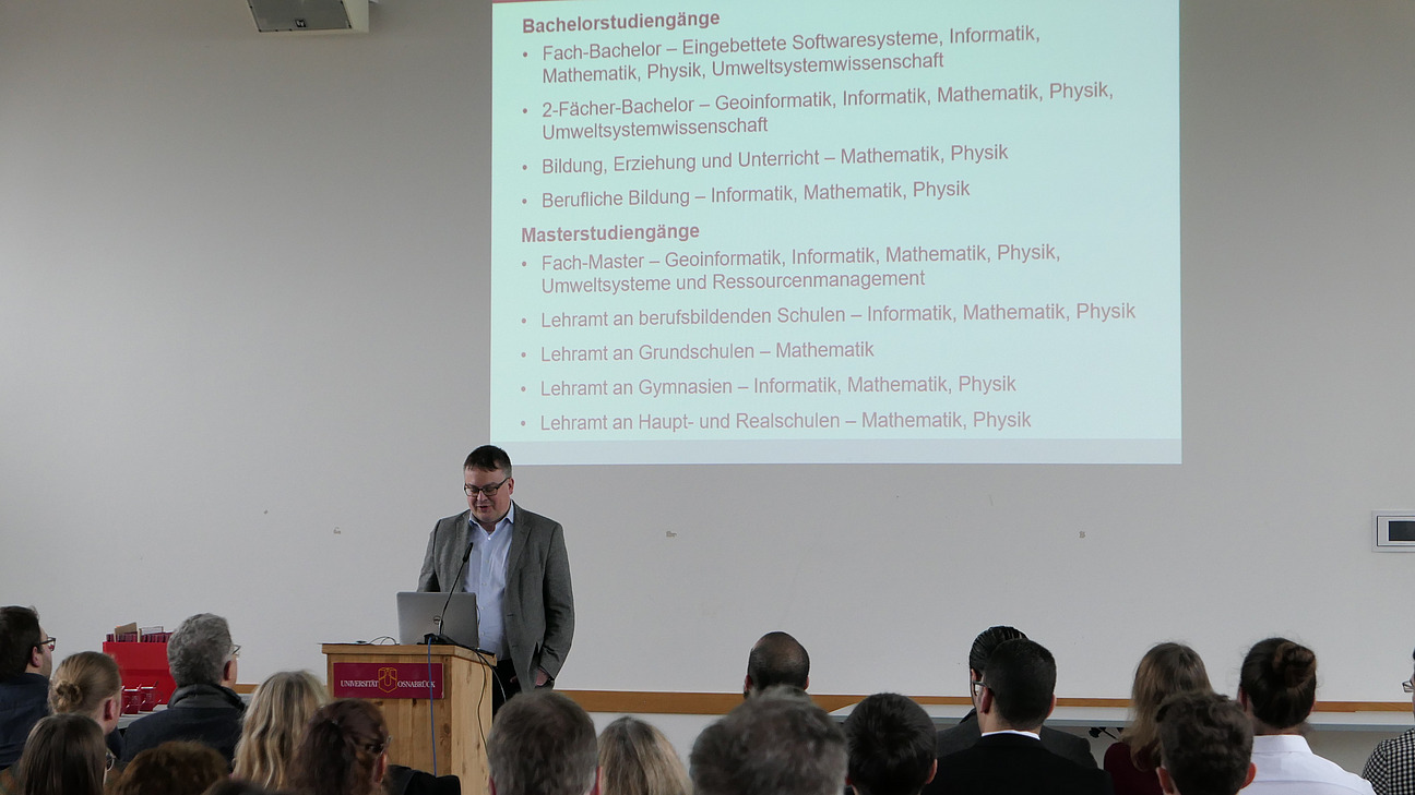 The dean is standing behind a lectern. The image of the projector can be seen behind him. 