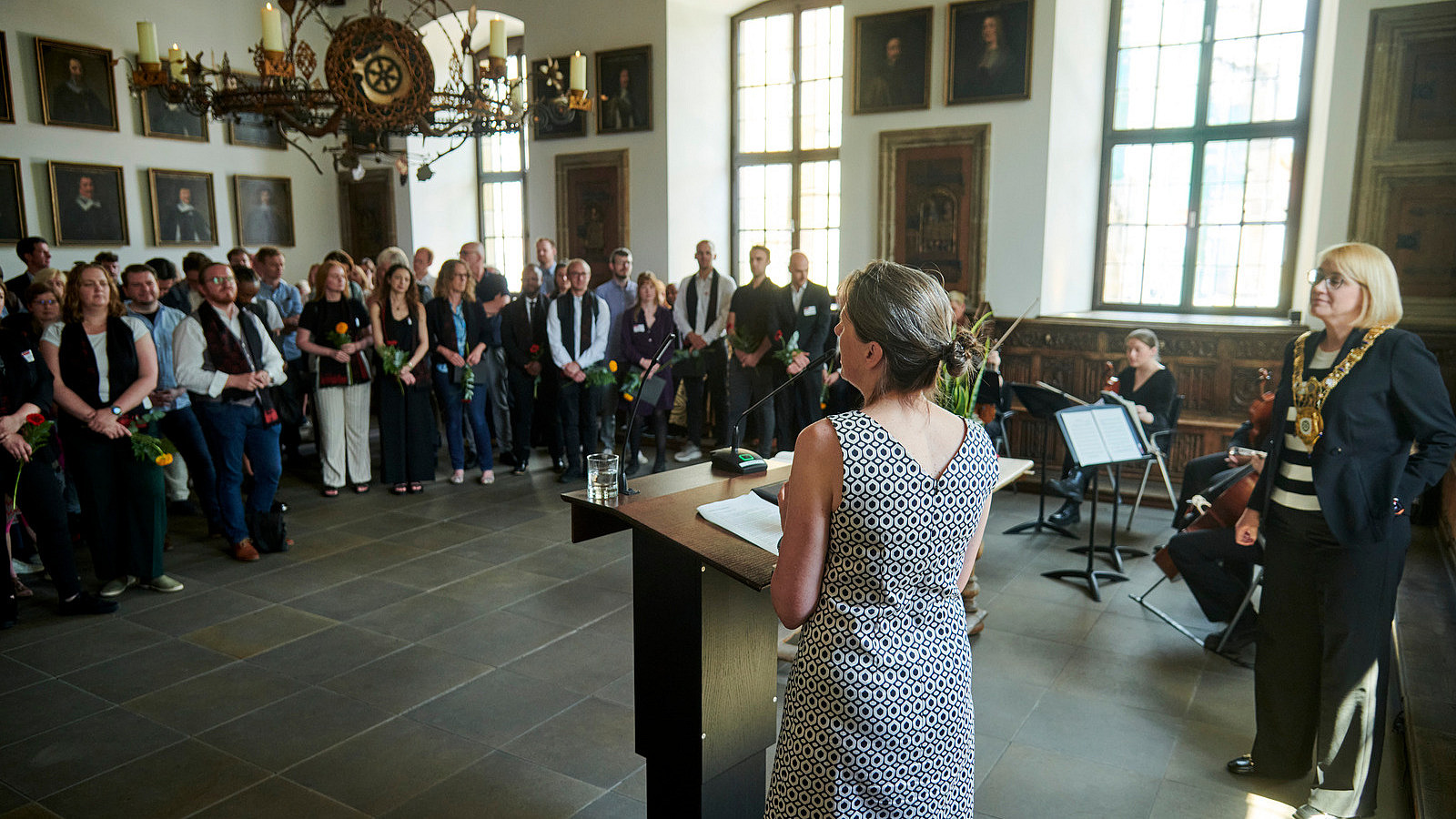 In a historical-looking room, a person stands at a lectern in the foreground. Numerous people are standing in front of her and looking at her.