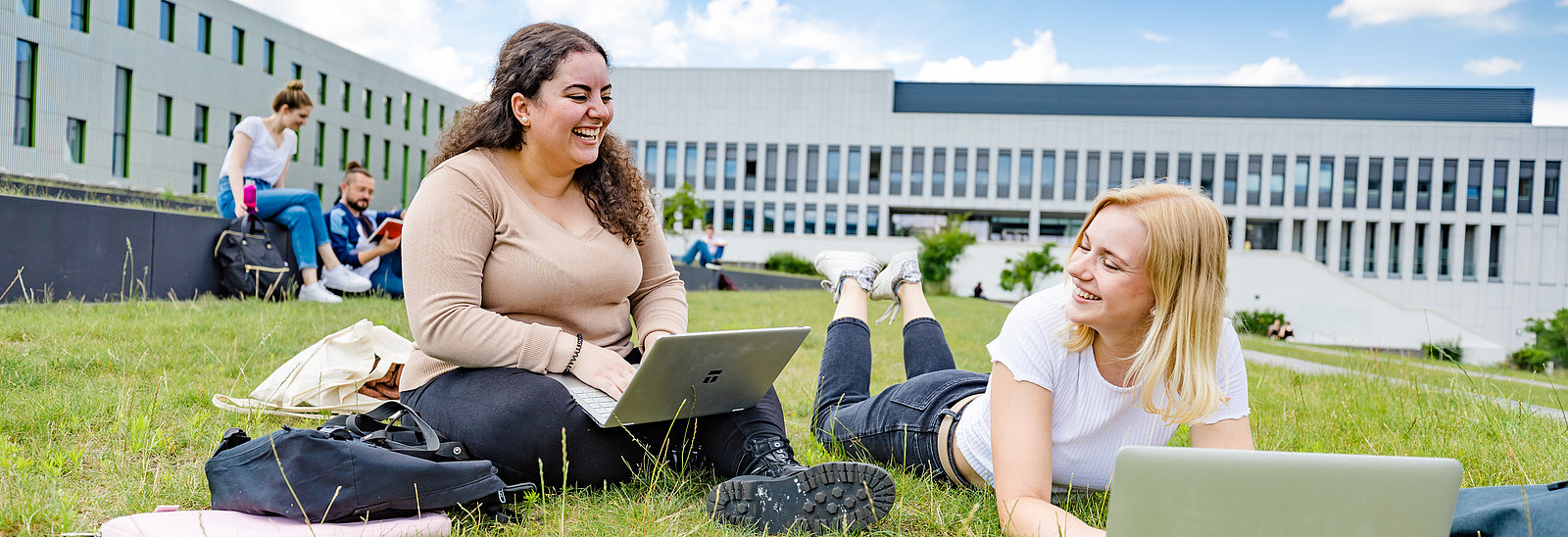 Zwei Studentinnen auf dem begrünten Platz neben der Bibliothek. Eine von ihnen sitzt, die andere liegt auf dem Rasen, beide haben Laptops vor sich