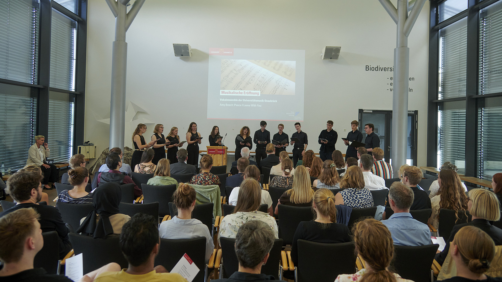 Viele Personen in einem Saal, Publikum von hinten, auf der Bühne stehen Musiker