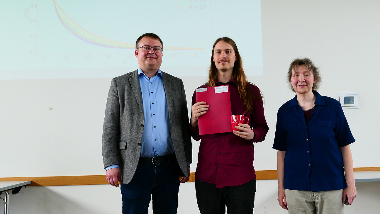Ein Student mit Studiendekanin und Dekan stehen nebeneinander. Der Student hat eine Mappe in der Hand