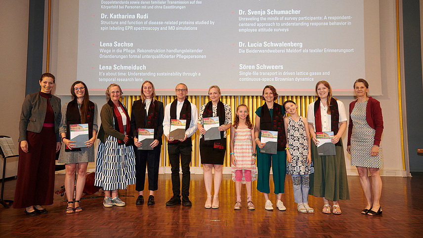 Several people stand on a stage, holding certificates in their hands and smiling into the camera