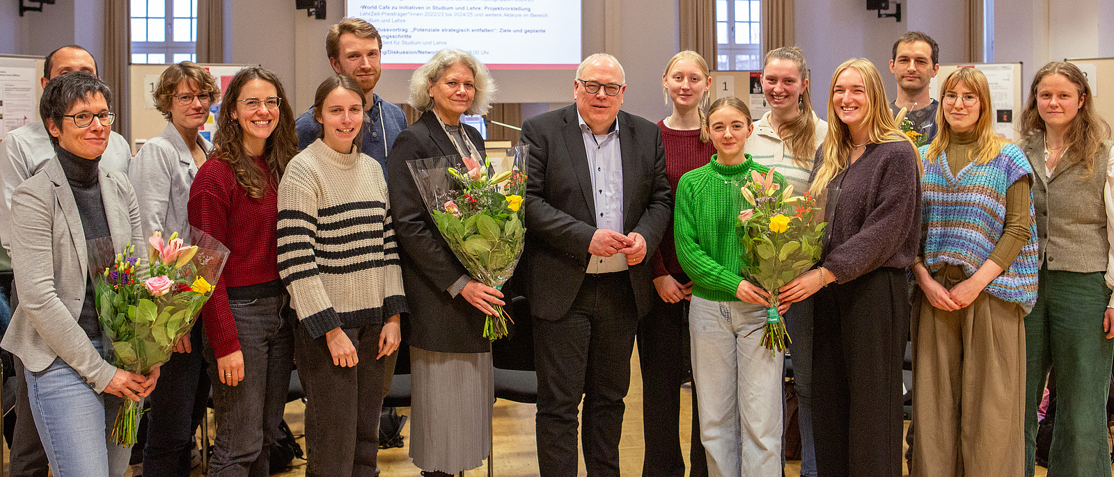 Men and women stand side by side, smiling and holding a bouquet of flowers.
