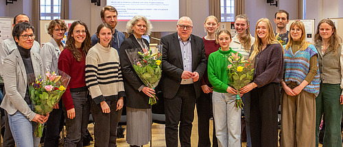 Men and women stand side by side, smiling and holding a bouquet of flowers.