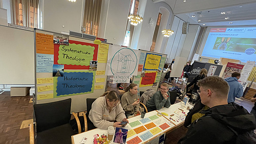 Mehrere Studierende sitzen eng beieinander an einem Tisch und arbeiten gemeinsam an Aufgaben. Sie beugen sich &uuml;ber Materialien und scheinen sich auszutauschen.  Im Hintergrund ist eine Wand mit vielen bunten Zetteln und Plakaten zu sehen. Auf einem gr&ouml;&szlig;eren Plakat steht &bdquo;Fachschaft Katholische Theologie&ldquo;. Die farbigen Zettel enthalten kurze Begriffe und Themen.   Weitere Personen sitzen im Hintergrund oder bewegen sich durch den Raum. Die Atmosph&auml;re ist lebendig und gemeinschaftlich.