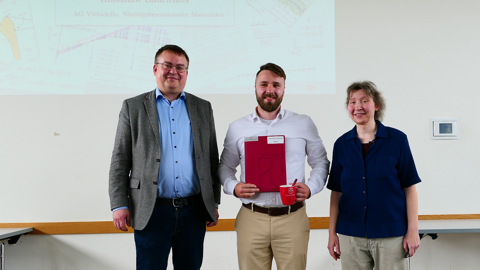 Ein Student mit Studiendekanin und Dekan stehen nebeneinander. Der Student hat eine Mappe in der Hand