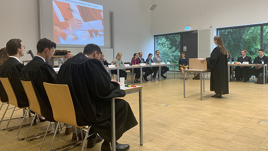 Frau in schwarzer Robe am Rednerpult vor einer "Richterbank" stehend, rechts und links Tische mit mehreren Personen in Roben