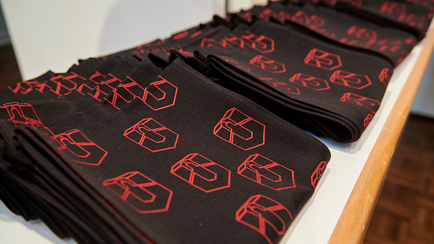 Close-up of a row of folded scarves. They are embroidered in black and red with a logo that is a combination of the letter U and the archway of a historic building.