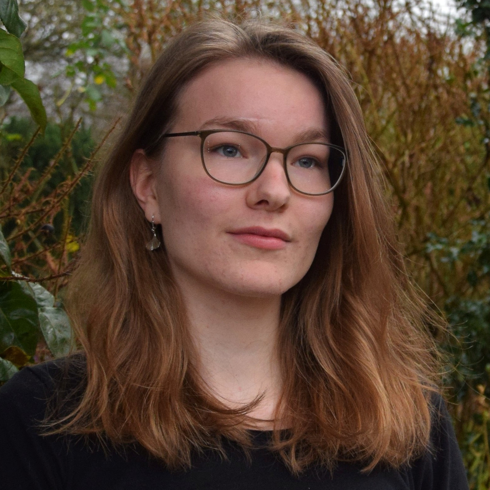 Portrait von einer jungen Frau mit braunen schulterlangen Haaren und brauner Brille. Sie hat ein schwarzes Langarmschirt an und im Hintergrund sind braune und gr&uuml;ne B&auml;ume und B&uuml;sche zu sehen.
