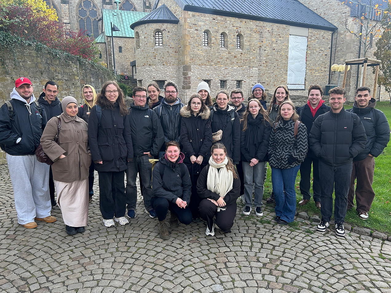 Das Bild zeigt eine Gruppe Menschen, die in Kamera lächeln. Im Hintergrund ist ein altes Haus aus Stein.