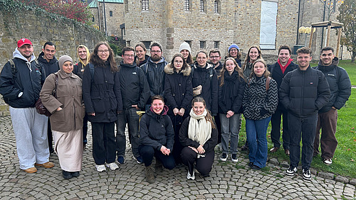 Das Bild zeigt eine Gruppe Menschen, die in Kamera lächeln. Im Hintergrund ist ein altes Haus aus Stein.