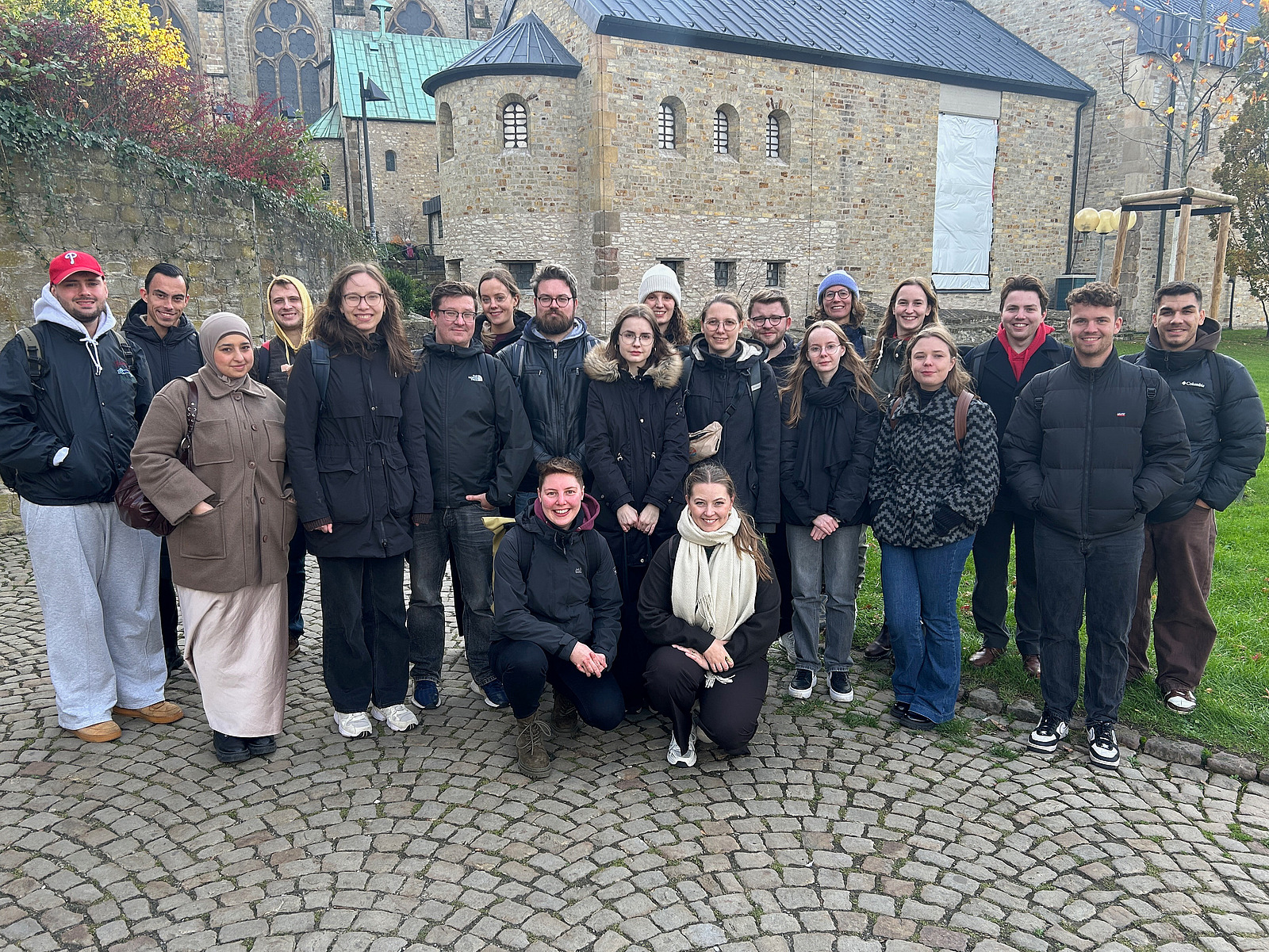 Das Bild zeigt eine Gruppe Menschen, die in Kamera lächeln. Im Hintergrund ist ein altes Haus aus Stein.