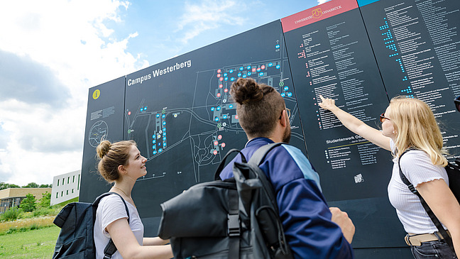 Ein paar Studierende zeigen mit dem Finger auf Geb&auml;ude auf einem gro&szlig;en Lageplan vom Westerberg
