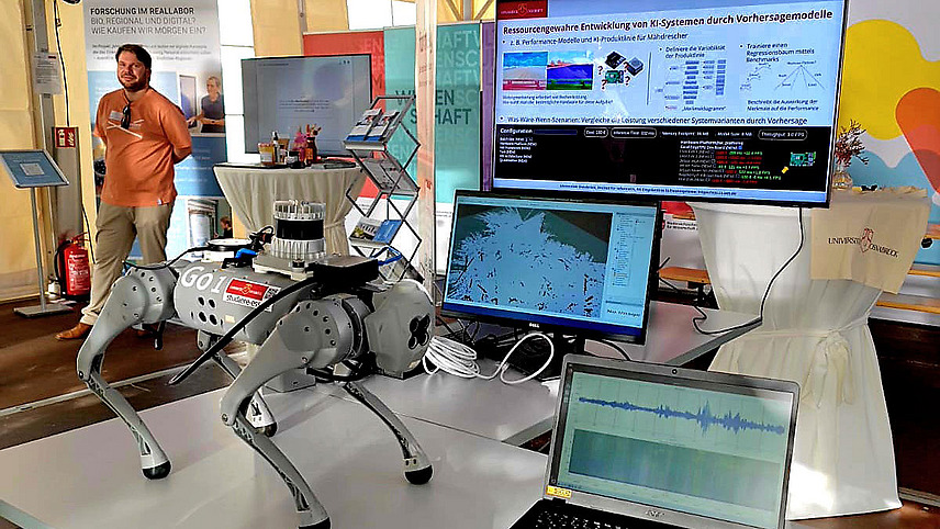 A dog-like robot stands in front of a booth with research posters.