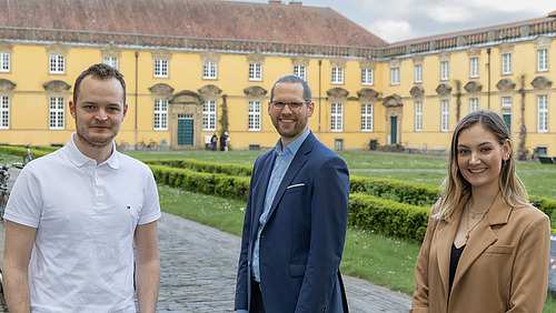 Drei Personen stehen im Vordergrund auf einem Weg und schauen in Richtung Kamera.  Hinter ihnen befindet sich ein gro&szlig;es, langgezogenes Geb&auml;ude mit gelber Fassade und vielen Fenstern. Davor liegt eine gepflegte Gr&uuml;nfl&auml;che mit niedrigen Hecken.  Im Hintergrund sind au&szlig;erdem B&auml;ume und ein Himmel mit Wolken zu sehen.