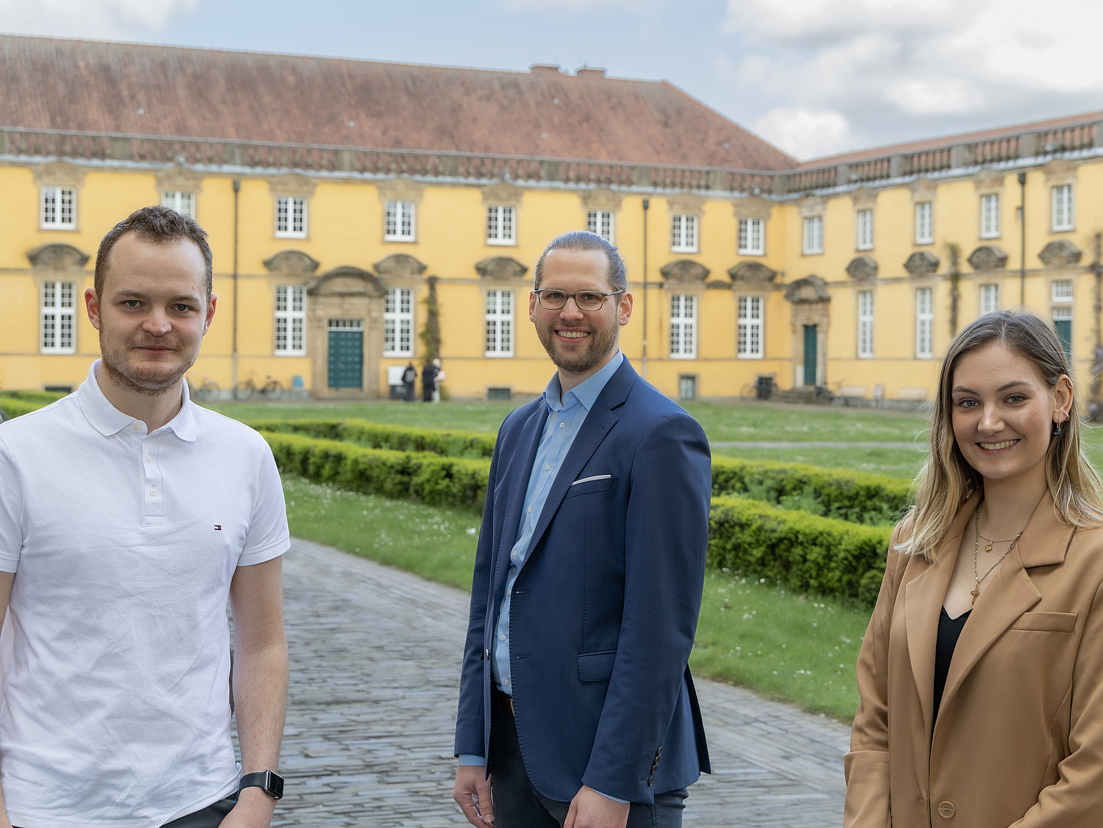Drei Personen stehen im Vordergrund auf einem Weg und schauen in Richtung Kamera.  Hinter ihnen befindet sich ein gro&szlig;es, langgezogenes Geb&auml;ude mit gelber Fassade und vielen Fenstern. Davor liegt eine gepflegte Gr&uuml;nfl&auml;che mit niedrigen Hecken.  Im Hintergrund sind au&szlig;erdem B&auml;ume und ein Himmel mit Wolken zu sehen.