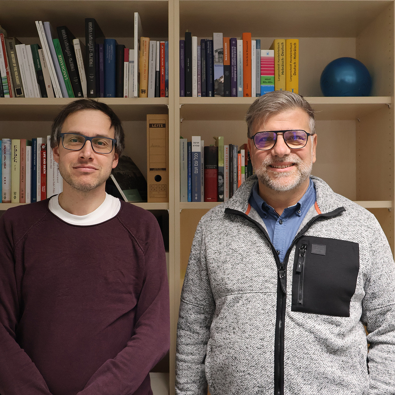 Zwei M&auml;nner mit Brillen stehen nebeneinander vor einem B&uuml;cherregal in einem B&uuml;ro und blicken in die Kamera.