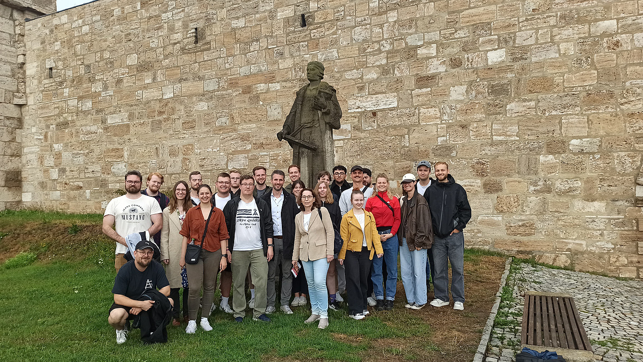 Das Bild zeigt eine Gruppe Menschen vor einer schwarzen Staute und einer Wand aus hellem Stein.