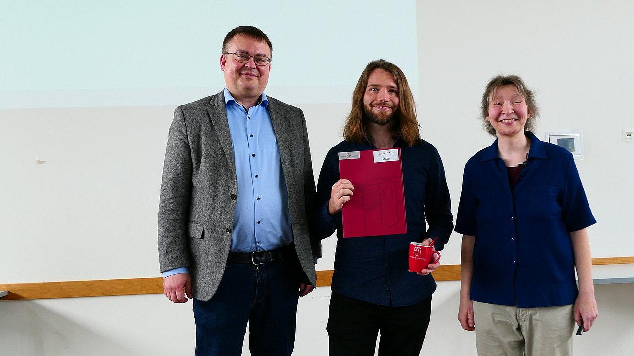 Ein Student mit Studiendekanin und Dekan stehen nebeneinander. Der Student hat eine Mappe in der Hand