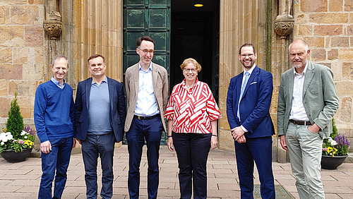 Sechs Personen stehen nebeneinander vor einem Geb&auml;ude mit steinerner Fassade und einer gro&szlig;en T&uuml;r im Hintergrund.  Die Personen sind in einer Reihe angeordnet und schauen in Richtung Kamera. Sie tragen unterschiedliche Kleidung, darunter Jacken, Hemden und ein auff&auml;lliges Oberteil mit Muster.  Links und rechts neben dem Eingang stehen Pflanzen in T&ouml;pfen.