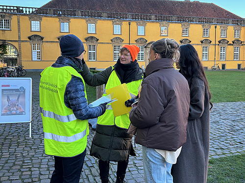 Two students in high-visibility vests stand in front of Osnabrück University and show two pupils the way. 