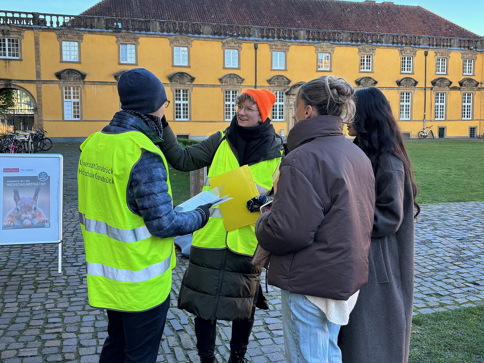 Two students in high-visibility vests stand in front of Osnabrück University and show two pupils the way. 