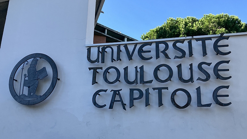 Logo und Schriftzug "Université de Toulouse Capitole" in blau auf weißer Wand vor blauem Himmel