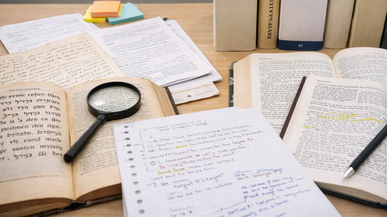 Ein Schreibtisch mit vielen aufgeschlagenen B&uuml;chern und Notizen. Mehrere dicke B&uuml;cher stehen auch aufrecht im Hintergrund, eines tr&auml;gt den Titel &bdquo;Exegese AT&ldquo;. Auf dem Tisch liegen offene Seiten mit markierten Textstellen. Daneben befinden sich handschriftliche Notizen auf losem Papier.  Eine Lupe liegt auf einem der B&uuml;cher, als w&uuml;rde jemand den Text genauer untersuchen. Ein Stift liegt bereit zum Schreiben. Insgesamt wirkt die Szene wie ein intensiver Arbeitsplatz zum Lesen, Lernen und Analysieren von Texten.