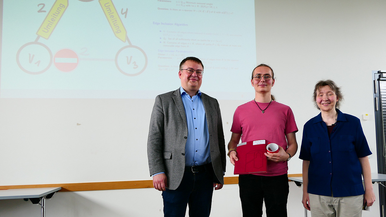 Ein Student mit Studiendekanin und Dekan stehen nebeneinander. Der Student hat eine Mappe in der Hand.