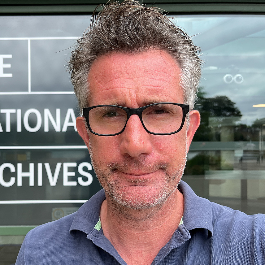 Ein Mann mit Brille und graumeliertem Haar steht vor einem Gebäude mit einem Schild, auf dem "The National Archives" steht. Er trägt ein blaues Polohemd und schaut mit leichtem Lächeln in die Kamera.