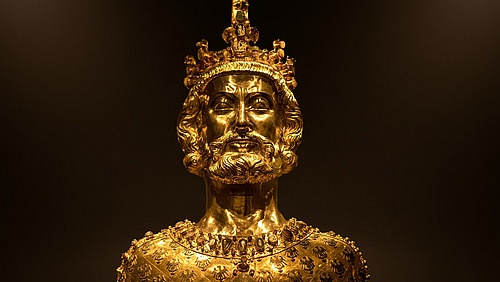 Eine goldfarbene B&uuml;ste zeigt den Kopf und Oberk&ouml;rper einer Person. Die Oberfl&auml;che ist mit vielen kleinen, unregelm&auml;&szlig;igen Elementen bedeckt.  Auf dem Kopf befindet sich ein kreuzf&ouml;rmiges Element. Das Gesicht ist nach vorne ausgerichtet.  Der Hintergrund ist dunkel, wodurch die goldene Figur deutlich hervorgehoben wird.