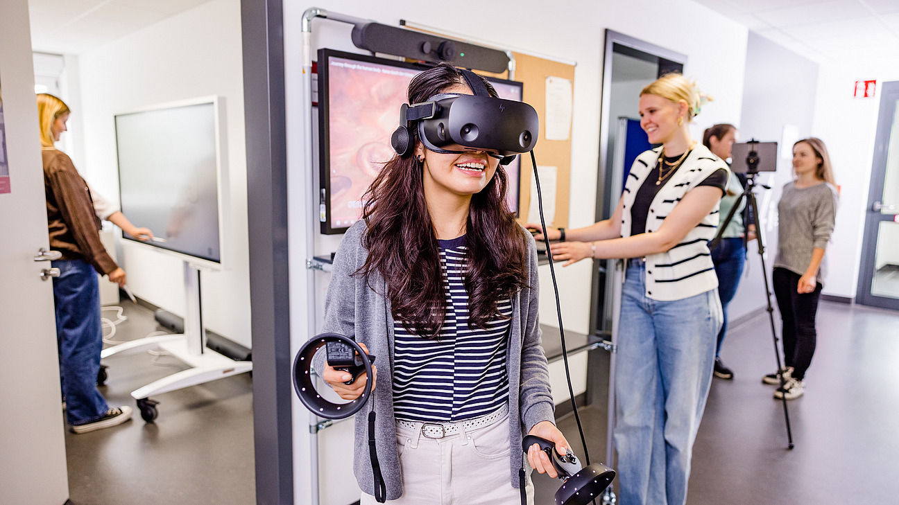 Eine Studierende tr&auml;gt eine Virtual Reality Brille. Im Hintergrund ist eine weitere Studierende zu sehen, welche die Simualtion &uuml;berwacht.