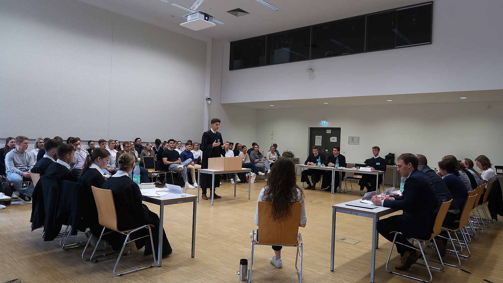 Junger Mann in schwarzer Robe hinter Rednerpult stehend, rechts und links Personen in Roben sitzend und davor Tischreihe mit "Richtern"