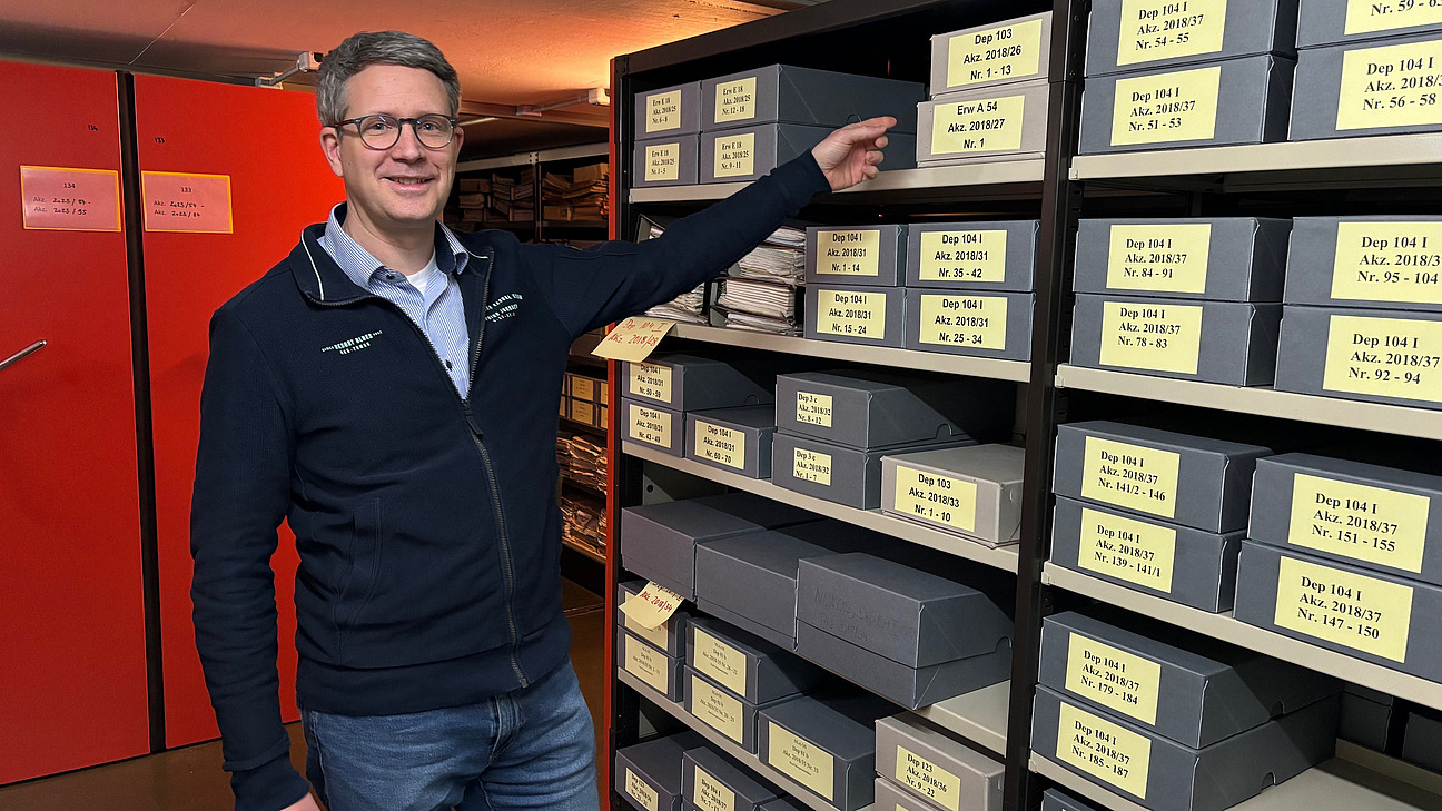 Ein Mann zeigt auf ein Regal voll Akten. 