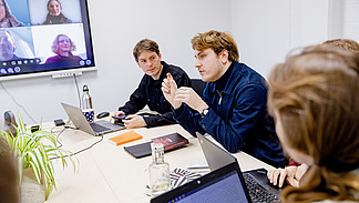 A group of people are holding a meeting at a conference table. One person is speaking while the others are sitting, some of them looking at laptops or notes. The other participants are shown on a screen in the background. 