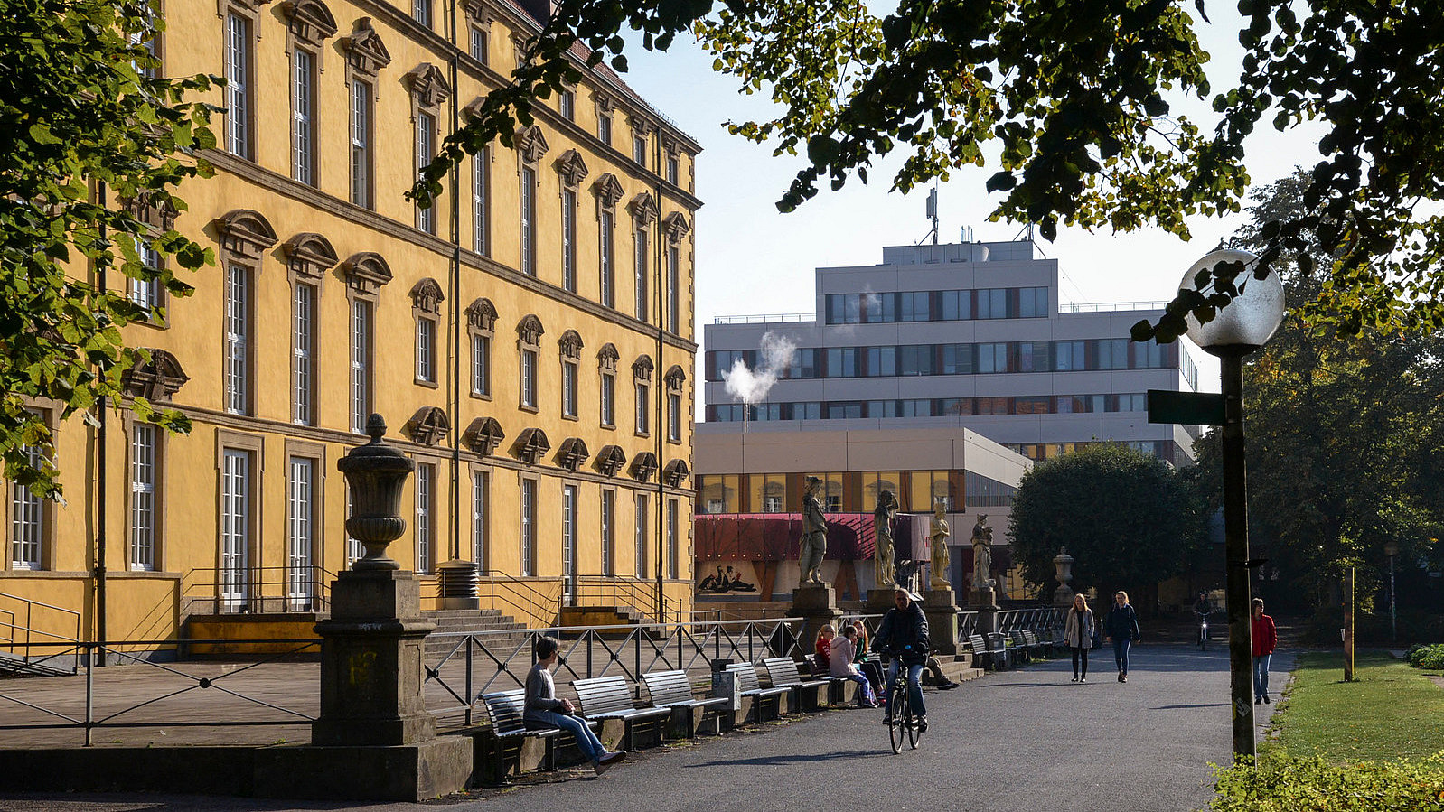 Seitliche Ansicht vom Gehweg zwischen Schloss und Schlossgarten. Beim Aufgang zur Terasse vor dem Schloss stehen vier Statuen aus Stein.