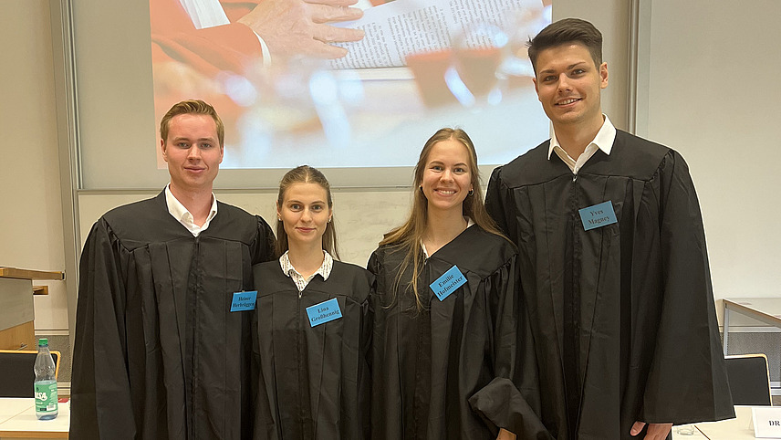 Zwei Frauen und zwei M&auml;nner in schwarzen Roben mit blauen Namensschildern in einer Reihe stehend