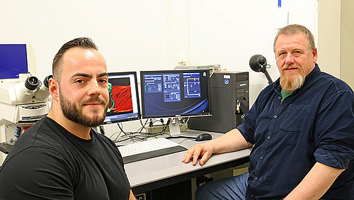 Two men sit at a desk with monitors and look into the camera.