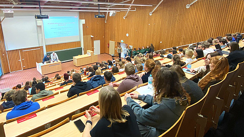 Blick von hinten in einen Hörsaal mit Studierenden, unten sind hinter einem Pult zwei Männer zu sehen.