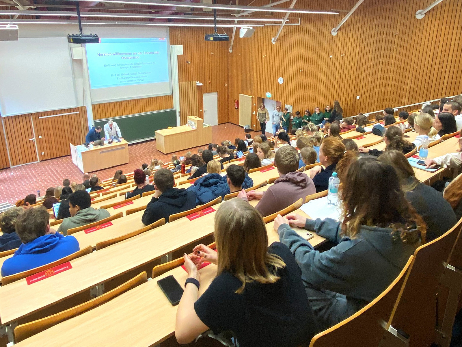 View from the back of a lecture hall with students; two men can be seen behind a desk at the bottom.