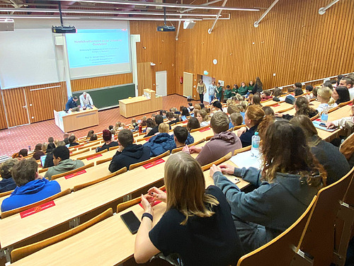Blick von hinten in einen Hörsaal mit Studierenden, unten sind hinter einem Pult zwei Männer zu sehen.